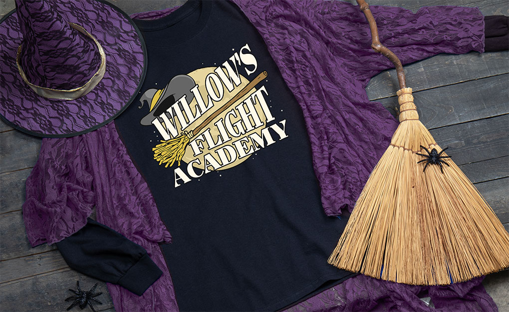 Your Name Flight Academy with a broom and witch's hat in front of a full moon on a long sleeve T shirt in a purple robe, next to a purple witch's hat and a gnarled broom.