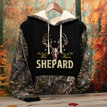 A871 American Hunter - A deer skull adorned with an overlaid American Flag, accompanied by the text Land Of The Free, Home Of The Brave, above Your Name, on a two tone camo hooded sweatshirt.