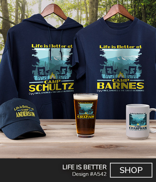 A542 | Life Is Better - Life Is Better At Camp Your Name, Enjoy Fires, S'mores, and The Great Outdoors, around trees, camp chairs, and a campfire by a lake with a mountain in the distance, on a navy T shirt, color printed pint glass, ceramic coffee mug, embroidered baseball hat, and a navy hoodie.