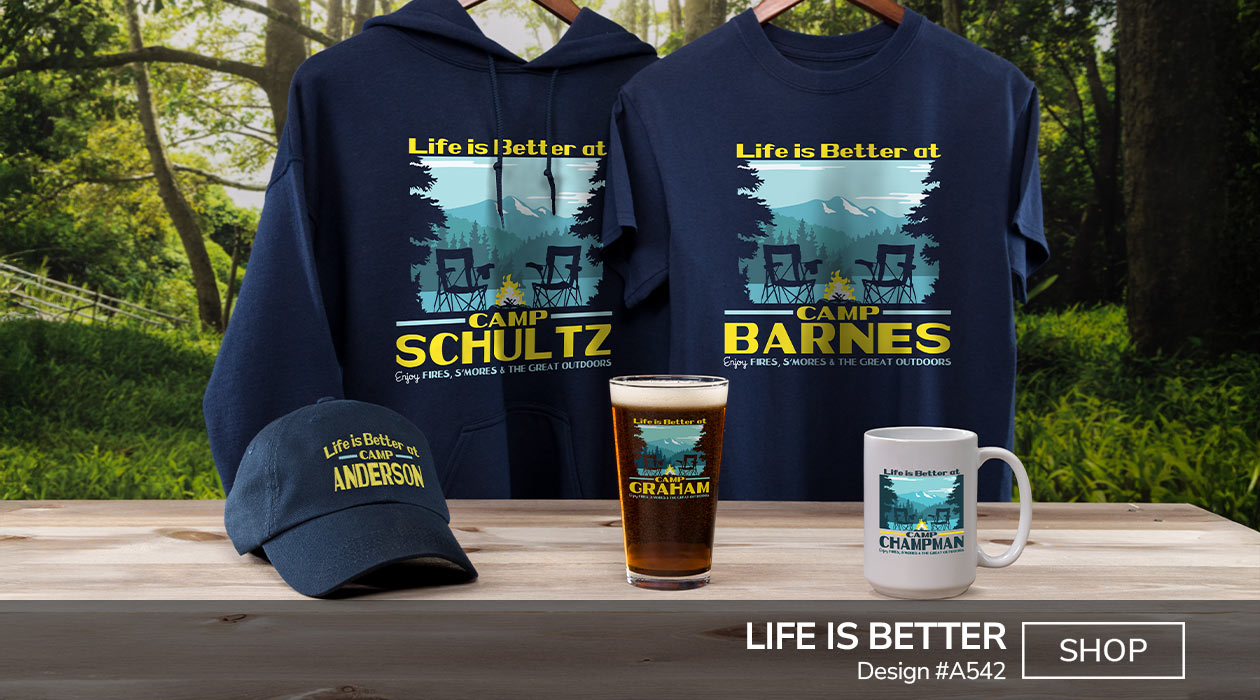 Design #A542 | Life Is Better - Life Is Better At Camp Your Name, Enjoy Fires, S'mores, and The Great Outdoors, around trees, camp chairs, and a campfire by a lake with a mountain in the distance, on a navy T shirt, color printed pint glass, ceramic coffee mug, and a navy hoodie.